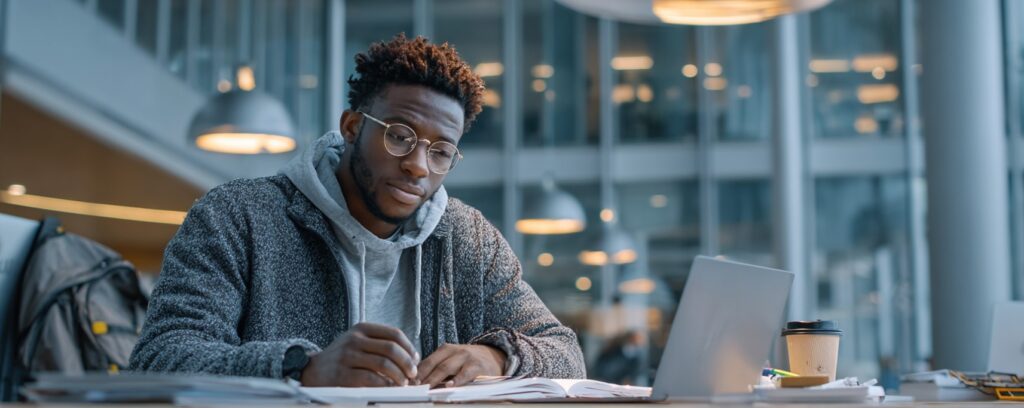 Young man studying at a desk with a laptop, notebook, and coffee in a modern, well-lit library or workspace.
